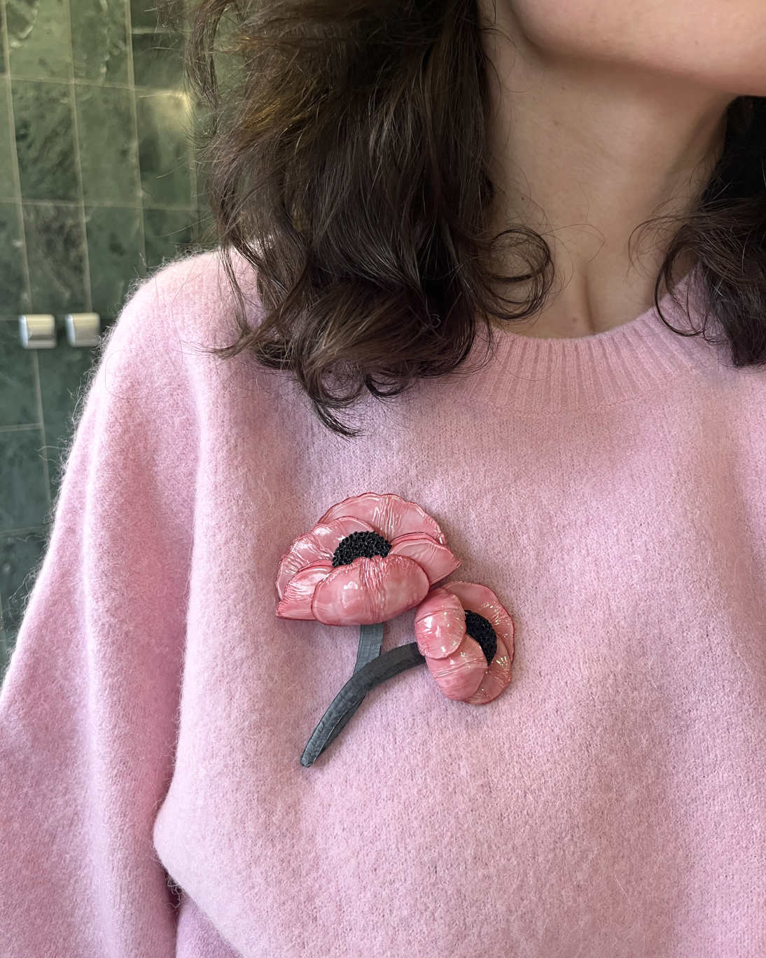 all-groups cette broche bijou unique rose Agnès représente de grandes fleurs qui se rejoignent en une tige. Elle est portée sur un pull rose par une jeune femme brune.